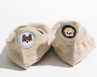 以下が含まれることがあります： 刺繍された動物の顔が付いたベージュのノースリーブソックスのペア。1つのソックスには茶色の犬の顔、もう1つのソックスには茶色の熊の顔があります。