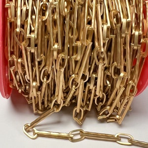 May include: A roll of gold-colored chain with a textured, hammered appearance. The chain links are rectangular and connected with a unique design. The chain is coiled on a pink spool, ready for use in jewelry making or crafts.