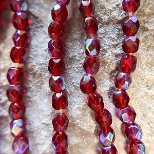 May include: Close-up of several strands of faceted, deep red beads. The beads have a multi-faceted cut, catching light and creating iridescent flashes of color. The beads are strung together, suggesting jewelry making or craft supplies.