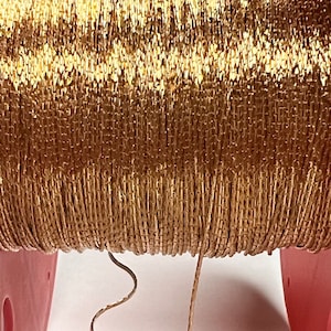 May include: A spool of gold metallic cord, wound tightly and showing the texture of the cord. The cord is a shiny gold color and has a slight sheen.
