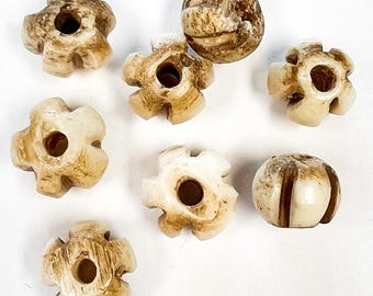May include: A collection of off-white, carved beads. Several are flower-shaped with a central hole, while others are spherical with vertical grooves. The beads have a natural, aged appearance with subtle brown markings.