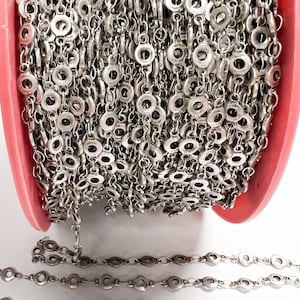 May include: Close-up of a roll of silver-colored chain with circular links. The chain is in a pink container, with a portion of the chain extending out. The chain is likely used for jewelry making or other craft projects.