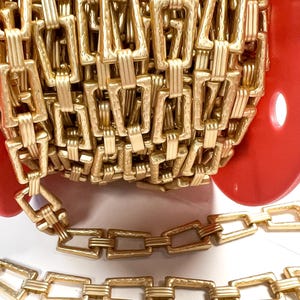 May include: Close-up of a gold-colored chain, with rectangular links. The chain is coiled on a red spool, with a portion of the chain extending out. The links have a textured surface.