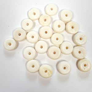 May include: A collection of off-white, donut-shaped beads with a central hole. The beads are smooth and appear to be made of a natural material. They are scattered on a white surface, suitable for jewelry making or crafts.