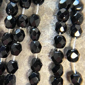 May include: A close-up of black faceted beads strung on white thread. The beads are round and have a shiny finish.