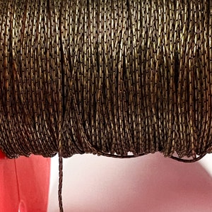 May include: Close-up of a spool of bronze-colored chain. The chain is tightly wound, creating a textured surface. The chain is made of small, rectangular links. The spool is partially visible, with a pink support structure in the background.