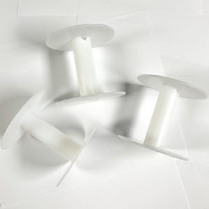 May include: Three white plastic spools with a disc-shaped top and bottom. The spools are arranged on a white surface, with the cylindrical center section connecting the two discs. The spools are made of a translucent white plastic.