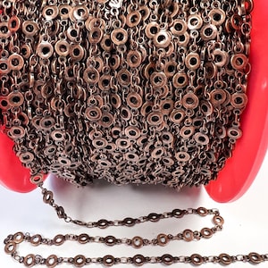 May include: Close-up of a roll of antique copper-colored chain. The chain is made of small, circular links with a central hole. The chain is displayed against a white background and a red surface.