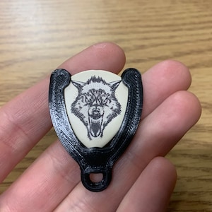 May include: A black 3D printed guitar pick holder with a wolf graphic on the pick. The holder has a loop for attaching to a belt or backpack.
