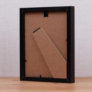 May include: A black picture frame with a brown backing and a built-in easel. The frame is rectangular and sits on a wooden surface. The back of the frame is visible, showing the easel and the backing material.
