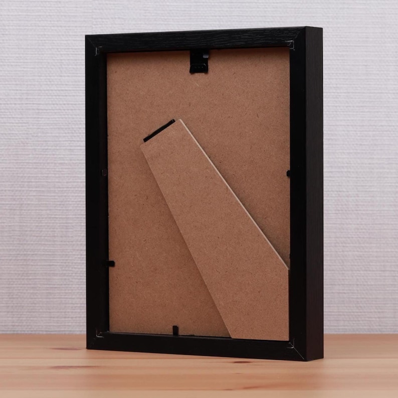 May include: A black picture frame with a brown backing and a built-in easel. The frame is rectangular and sits on a wooden surface. The back of the frame is visible, showing the easel and the backing material.