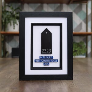 May include: Framed black police badge with white matting. The badge is black with the number 2323 and the text "PC TILEHURST WEST YORKSHIRE POLICE 2026" in white and blue. The frame is black.