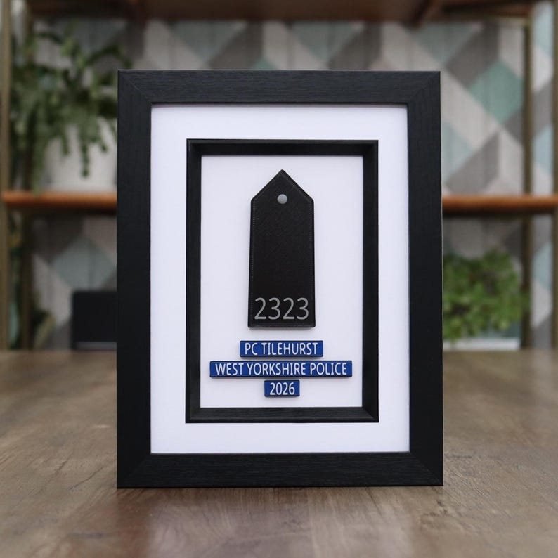 May include: Framed black police badge with white matting. The badge is black with the number 2323 and the text "PC TILEHURST WEST YORKSHIRE POLICE 2026" in white and blue. The frame is black.