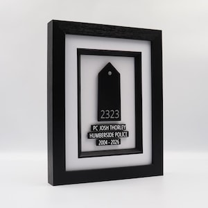 May include: A framed black plaque with a white mat. The plaque features a black tag-shaped design with the number "2323" and the text "PC JOSH THORLEY HUMBERSIDE POLICE 2004-2026" in white. The frame is black.