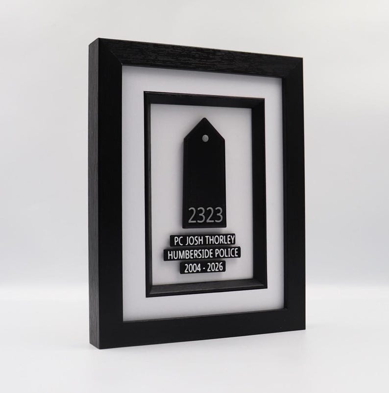 May include: A framed black plaque with a white mat. The plaque features a black tag-shaped design with the number "2323" and the text "PC JOSH THORLEY HUMBERSIDE POLICE 2004-2026" in white. The frame is black.