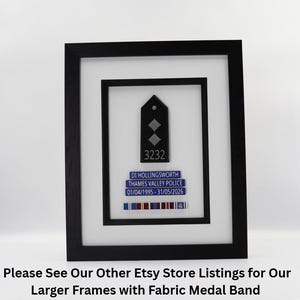 May include: Framed police badge display. The black frame surrounds a white matting, showcasing a black badge with two silver diamonds and the number 3232. Below, a blue nameplate reads "DI HOLLINGSWORTH, THAMES VALLEY POLICE." A ribbon bar with colored stripes completes the display.