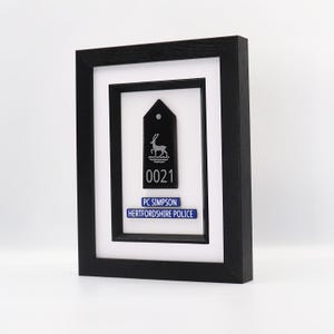May include: Framed black police badge with a deer emblem, the number 0021, and the text "PC SIMPSON HERTFORDSHIRE POLICE" on a blue background. The frame is black and the background is white.