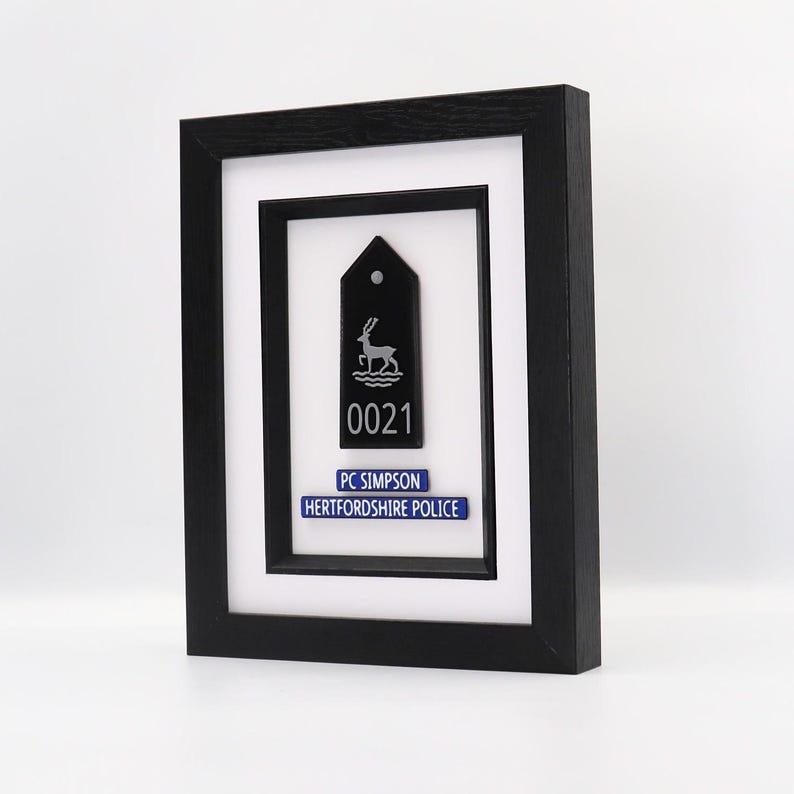 May include: Framed black police badge with a deer emblem, the number 0021, and the text "PC SIMPSON HERTFORDSHIRE POLICE" on a blue background. The frame is black and the background is white.