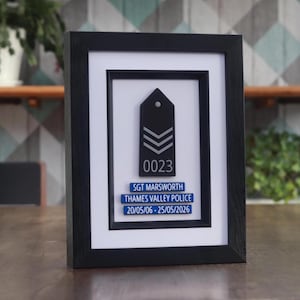 May include: Framed black and white police sergeant rank insignia with the number "0023" and the text "SGT MARSWORTH THAMES VALLEY POLICE 20/05/06 - 25/05/2026". The frame is black and the background is white.