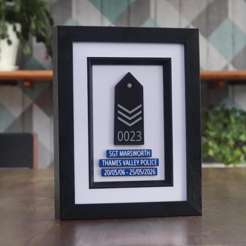May include: Framed black and white police sergeant rank insignia with the number "0023" and the text "SGT MARSWORTH THAMES VALLEY POLICE 20/05/06 - 25/05/2026". The frame is black and the background is white.