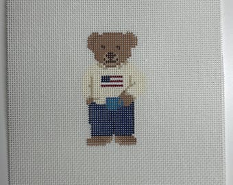 May include: Needlepoint design of a brown teddy bear wearing a cream-colored sweater with a red, white, and blue American flag design. The bear is wearing blue pants and brown shoes. The background is a white canvas.