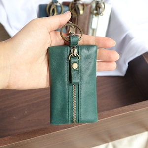 May include: A green leather key chain with a zipper closure and a metal key ring. The key chain is held in a hand.