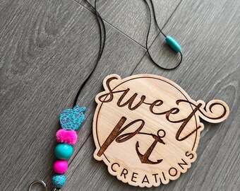 Puede incluir: Un cordón con un diseño de cuentas turquesas y rosas, un cierre plateado y un dije turquesa. El cordón tiene un cordón negro y está junto a un letrero de madera que dice "Sweet Pit Creations".