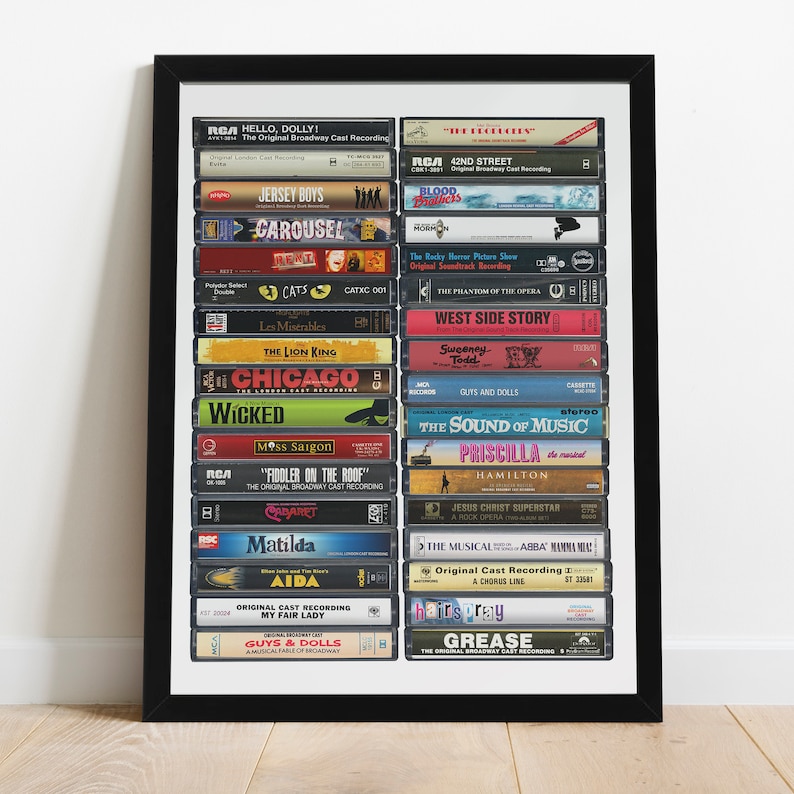 May include: A framed print of a collection of VHS tapes featuring the original Broadway cast recordings of popular musicals. The tapes are arranged vertically in a black frame and include titles like "Hello, Dolly!" "Jersey Boys", "Cats", "Les Miserables", "The Lion King", "Chicago", "Wicked", "Miss Saigon", "Fiddler on the Roof", "Matilda", "Aida", "My Fair Lady", "Guys & Dolls", "42nd Street", "Blood Brothers", "The Rocky Horror Picture Show", "The Phantom of the Opera", "West Side Story", "Sweeney Todd", "Guys and Dolls", "The Sound of Music", "Priscilla", "Hamilton", "Jesus Christ Superstar", "The Musical ABBA Mamma Mia", "A Chorus Line", "Hairspray", and "Grease".
