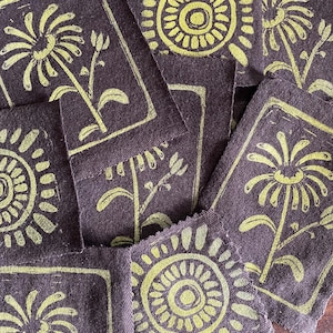 以下が含まれることがあります： 濃い茶色の布の正方形のコレクションで、黄緑色の花と太陽の模様が繰り返されています。 花の要素はブロックプリントされており、素朴で手作りの美しさを生み出しています。