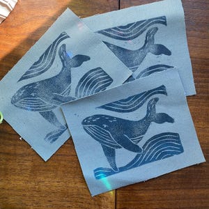 Pode incluir: Quatro quadrados de tecido com um desenho de baleia azul. As baleias são retratadas saltando para fora da água com ondas ao fundo. Os quadrados são de cor azul clara e estão dispostos sobre uma superfície de madeira.