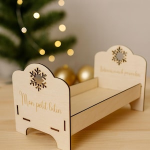 Può includere: Un piccolo letto in legno chiaro, decorato con intagli a forma di fiocchi di neve e la scritta "Mon petit lutin". Questo letto in miniatura, dal design semplice ed elegante, è probabilmente un oggetto decorativo per le feste.