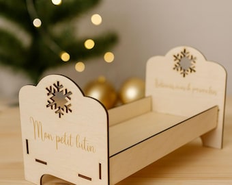 Peut inclure: Un petit lit en bois clair, orné de découpes en forme de flocons de neige et de l'inscription "Mon petit lutin". Ce lit miniature, de conception simple et élégante, est probablement un objet décoratif pour les fêtes.