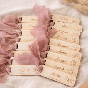 Può includere: Tag di nome in legno con nomi in elegante scrittura, accentati da un motivo a margherita. Nastri rosa polvere sono attaccati ai tag. I tag sono disposti su una superficie color crema.
