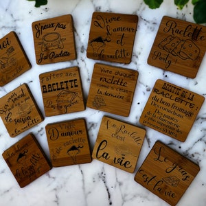 Op de afbeelding: Een verzameling vierkante bamboe onderzetters met gegraveerde tekst en illustraties. De onderzetters bevatten zinnen als "Raclette Party!" en "La vie". Geschikt voor gebruik in een keuken of eetkamer.