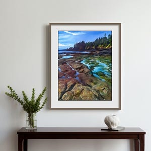 Op de afbeelding: Een ingelijste landschapskunstafdruk met een kustlandschap met kleurrijke rotsen, water en bomen. Het kunstwerk hangt boven een donkere houten console met een vaas met groen en een decoratief object. De kamer heeft witte muren en een raam.