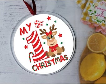 Puede incluir: Adorno navideño redondo con el texto "MY 1st CHRISTMAS". El adorno presenta un reno de dibujos animados, un bastón de caramelo y estrellas en rojo, verde y blanco. Se adjunta una cinta roja para colgar.