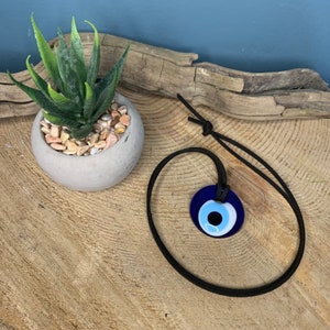 May include: A blue evil eye charm necklace with a black cord. The charm is round and has a white and blue eye design.
