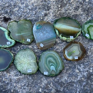 May include: Eight agate slices with green and white patterns. The slices are arranged in a semi-circle on a grey, textured surface.