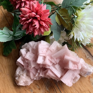 Peut inclure: Un amas de cristaux rose clair à la structure géométrique. Posé sur une surface en bois, l'image présente également un bouquet de fleurs roses et blanches en arrière-plan.
