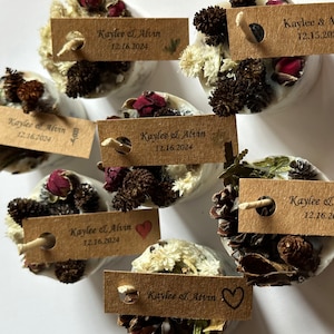 May include: A set of eight small, round, white candles with brown, white, and red dried flowers and greenery. Each candle has a small, brown paper tag with the text "Kaylee & Alvin" and a date.