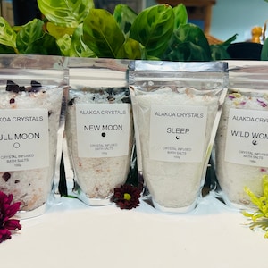 May include: Four clear plastic pouches filled with white bath salts. Each pouch has a label with the text "Alakoa Crystals" and a different moon phase: Full Moon, New Moon, Sleep, and Wild Woman. Each pouch also includes the text "Crystal Infused Bath Salts 150g".