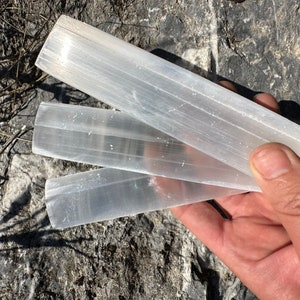 May include: Three selenite wands, also known as satin spar, are stacked on top of each other. The wands are white and translucent, with a smooth, polished surface. The wands are approximately 10 cm long and 2.5 cm wide.