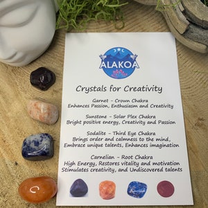 May include: A set of four crystals for creativity, including garnet, sunstone, sodalite, and carnelian. Each crystal is labelled with its corresponding chakra and benefits. The crystals are arranged on a wooden surface with green moss.