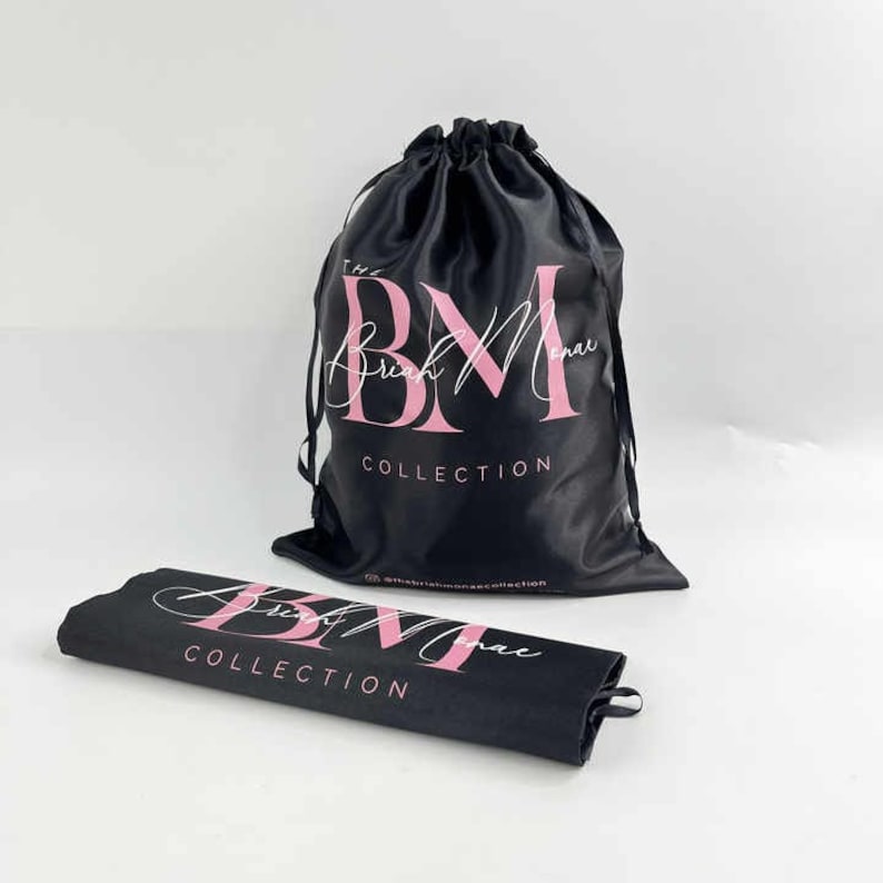 May include: Black satin drawstring bag with the text "The Briah Monae Collection" in pink lettering. A matching black fabric rectangle with the same text is also shown.