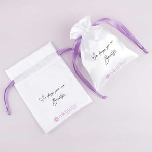 May include: Two white satin drawstring bags with purple ribbon ties. Both bags have the text "We design your own Beauty" and the logo "mktg industry" printed on them.