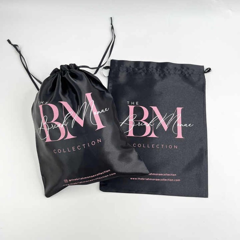 May include: Two black satin drawstring bags with the text "The Brush Monae Collection" and the logo "BM" in pink lettering.