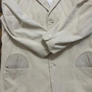 May include: A light beige corduroy blazer with a diamond-quilted upper chest. The blazer features two front pockets, a notched lapel, and three buttons. The long sleeves have buttoned cuffs. The interior lining is white.