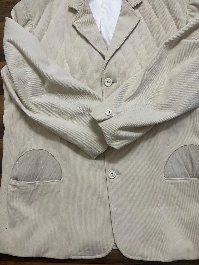May include: A light beige corduroy blazer with a diamond-quilted upper chest. The blazer features two front pockets, a notched lapel, and three buttons. The long sleeves have buttoned cuffs. The interior lining is white.