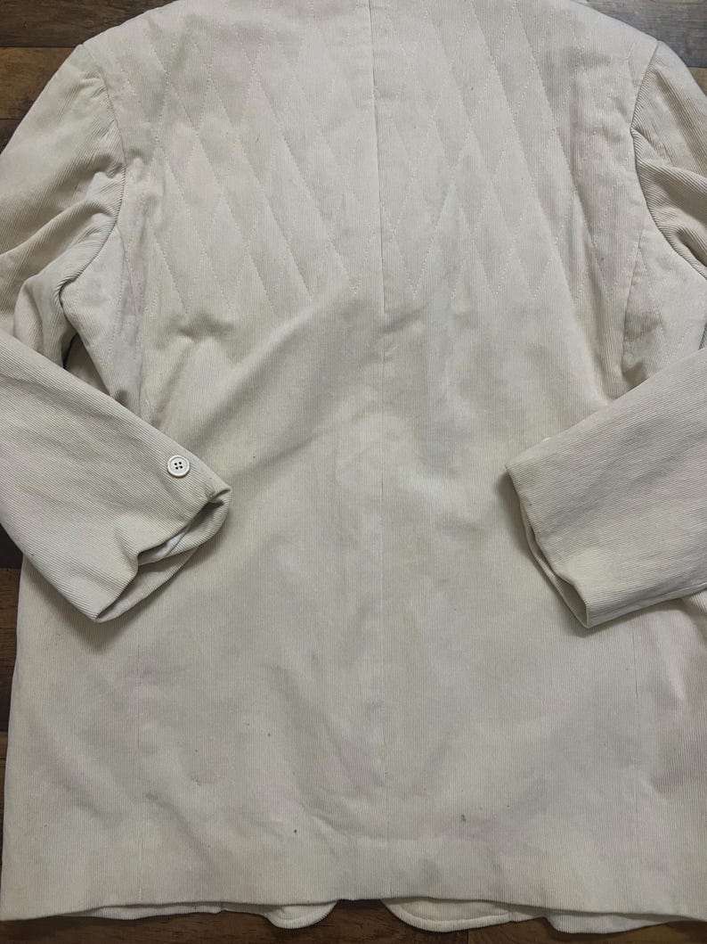 May include: A light beige corduroy blazer. The back panel features a quilted design. The blazer has long sleeves with buttoned cuffs and a notched lapel. The fabric has a vertical ribbed texture.