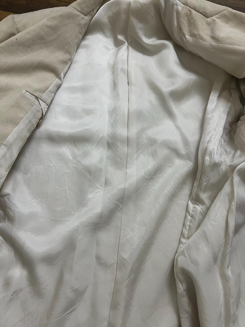 May include: Beige corduroy blazer with a white satin lining. The interior of the jacket is visible, showcasing the smooth, shiny texture of the lining. The blazer's exterior has a ribbed texture.